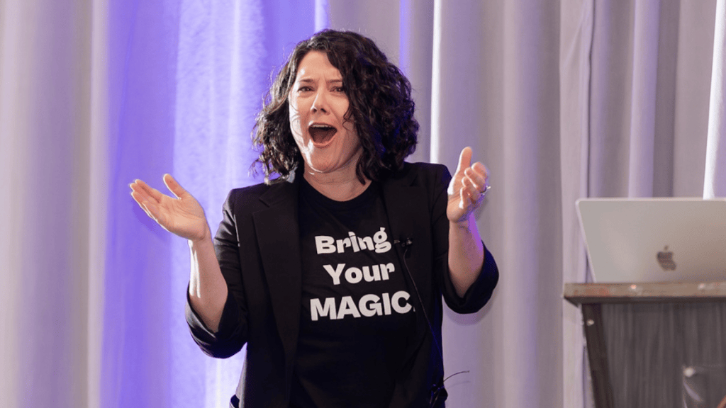 Su Zangara speaking on stage in a “Bring Your Magic” t-shirt